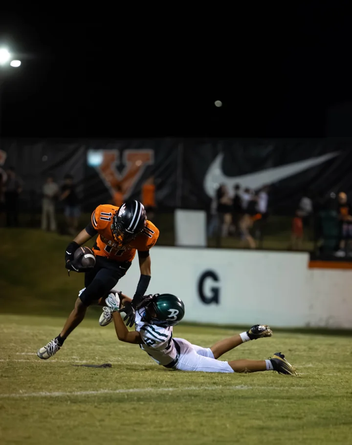 A football player in an orange uniform runs with the ball as a defender reaches from the ground.