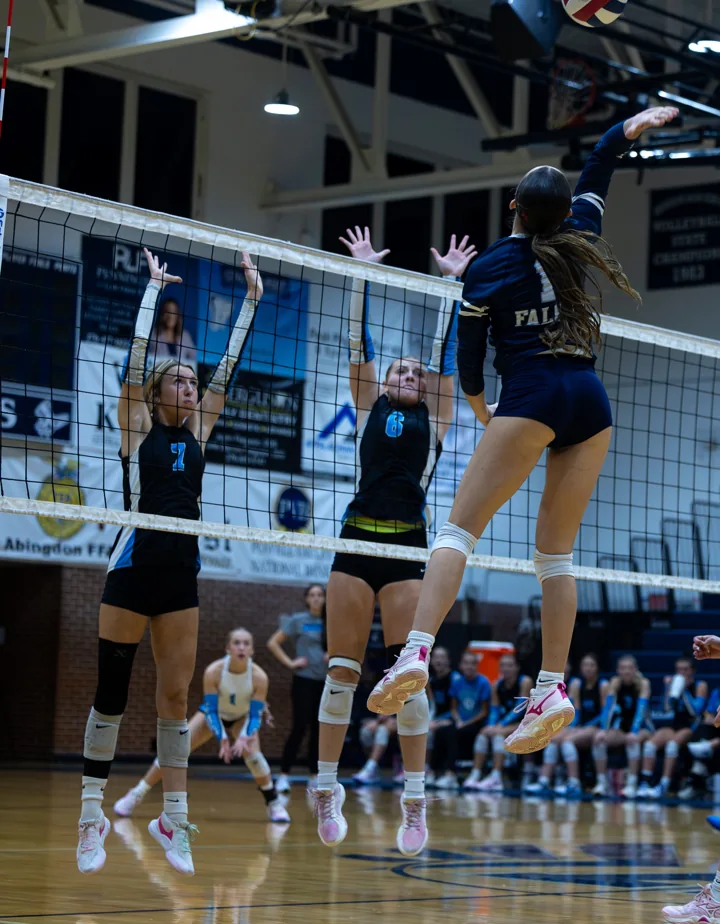 A volleyball player leaps to attack the ball while defenders prepare to block at the net.