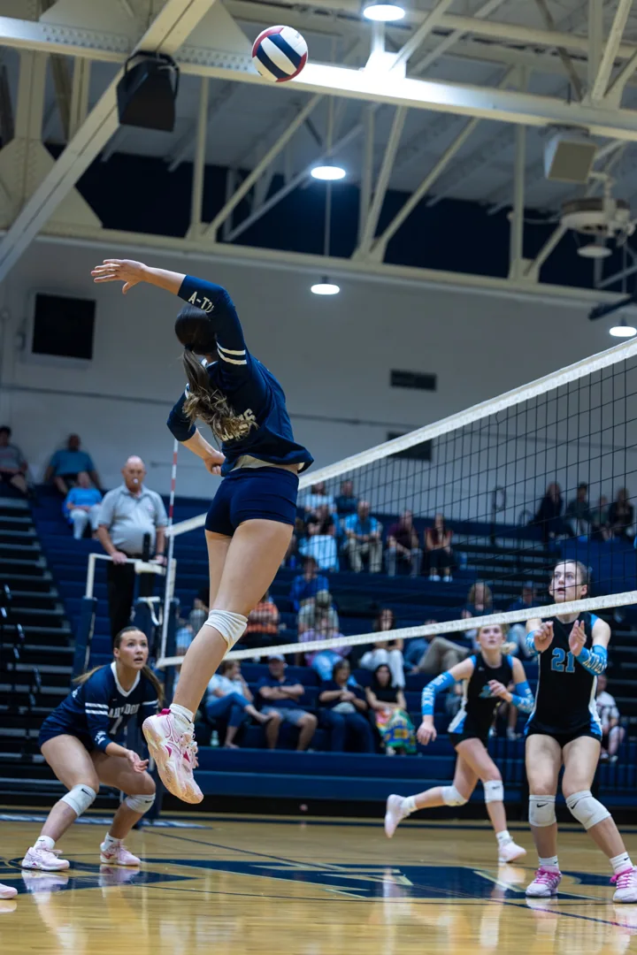 A volleyball player in navy jumps high at the net with the ball above her hitting arm.