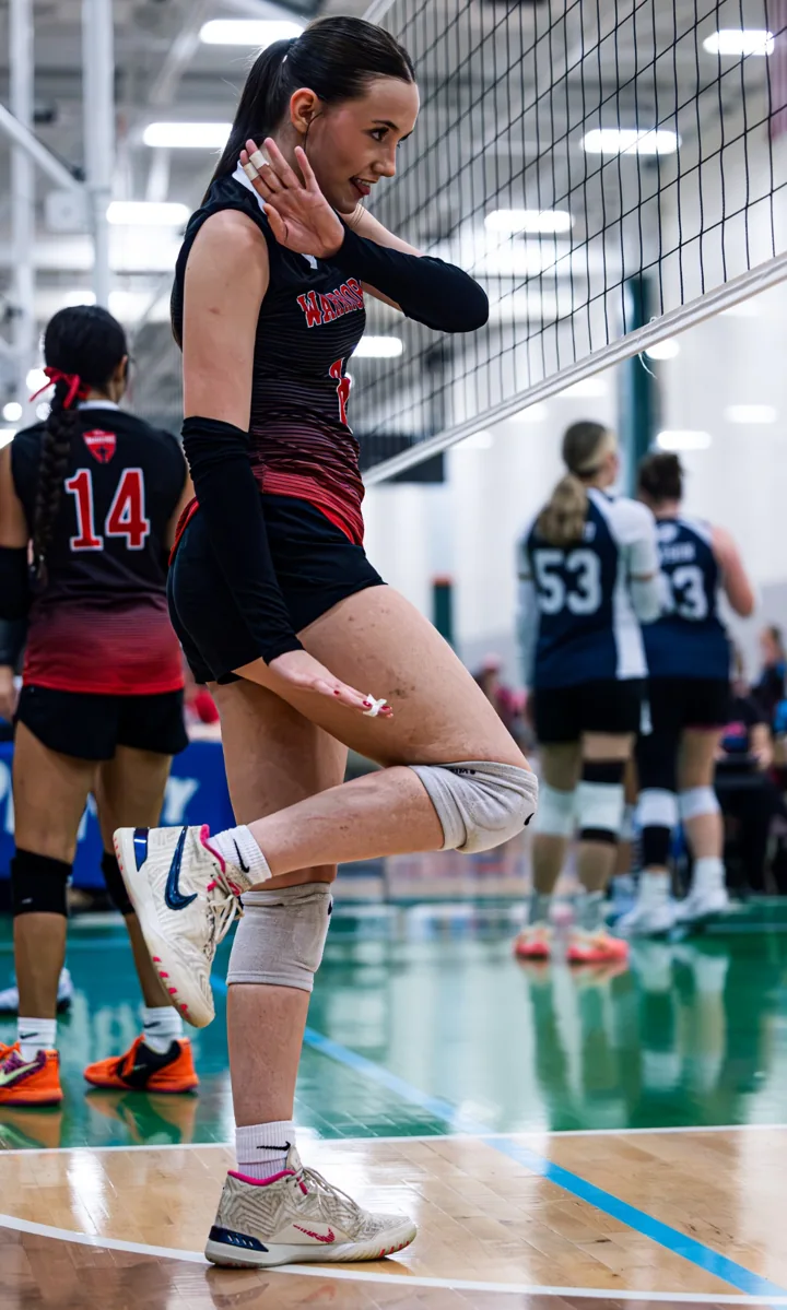 A volleyball player in red and black adjusts her stance between points near the net.