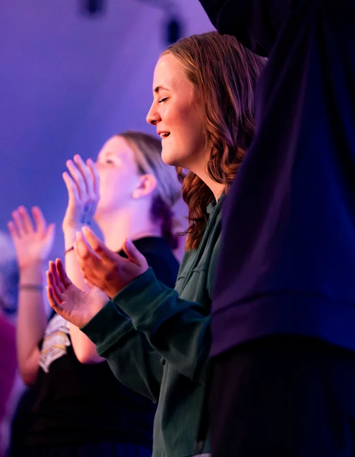 People raise their hands and sing together under purple stage lighting.