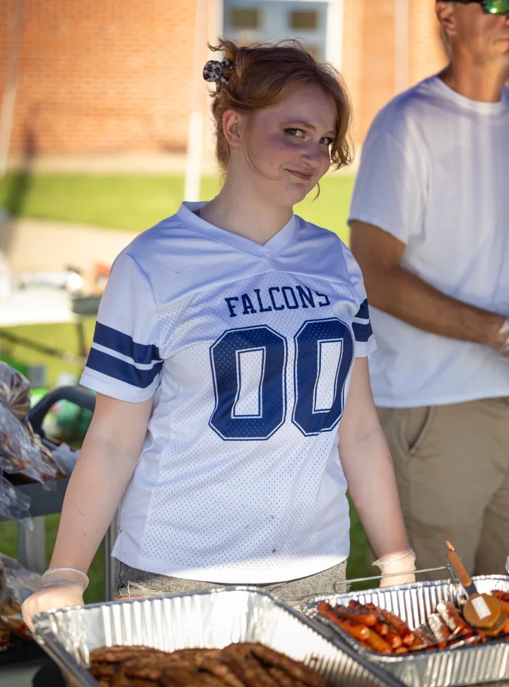 A volunteer in a football-style jersey stands beside trays of food at an outdoor gathering.
