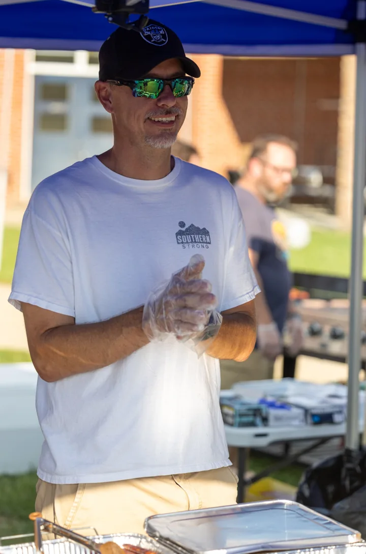 A volunteer stands behind food trays under a canopy at a community gathering.
