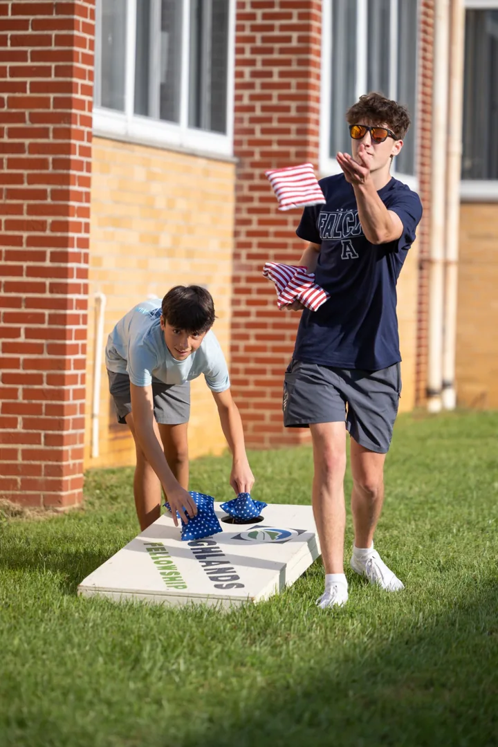 A young man plays a beanbag toss game on a lawn beside a brick building.