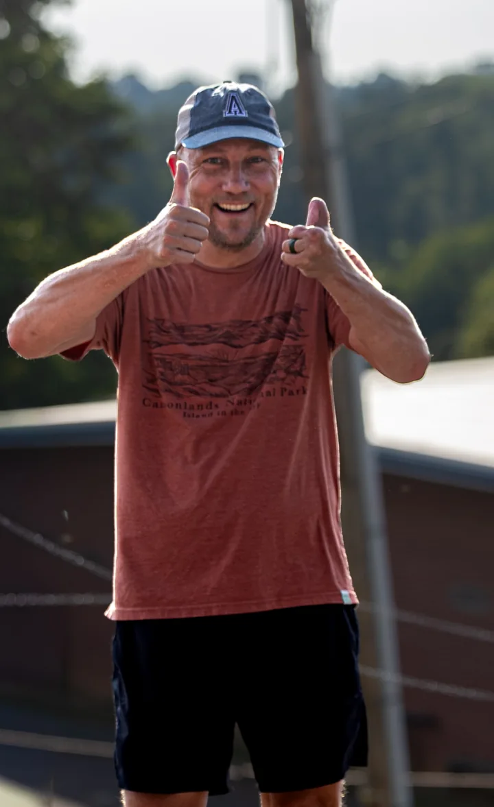 A smiling man in a cap gives a playful hand gesture outdoors.