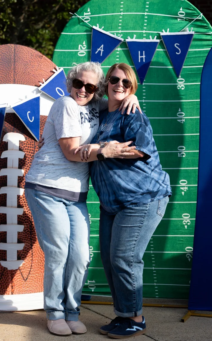 Two smiling people embrace in front of a football-themed backdrop.