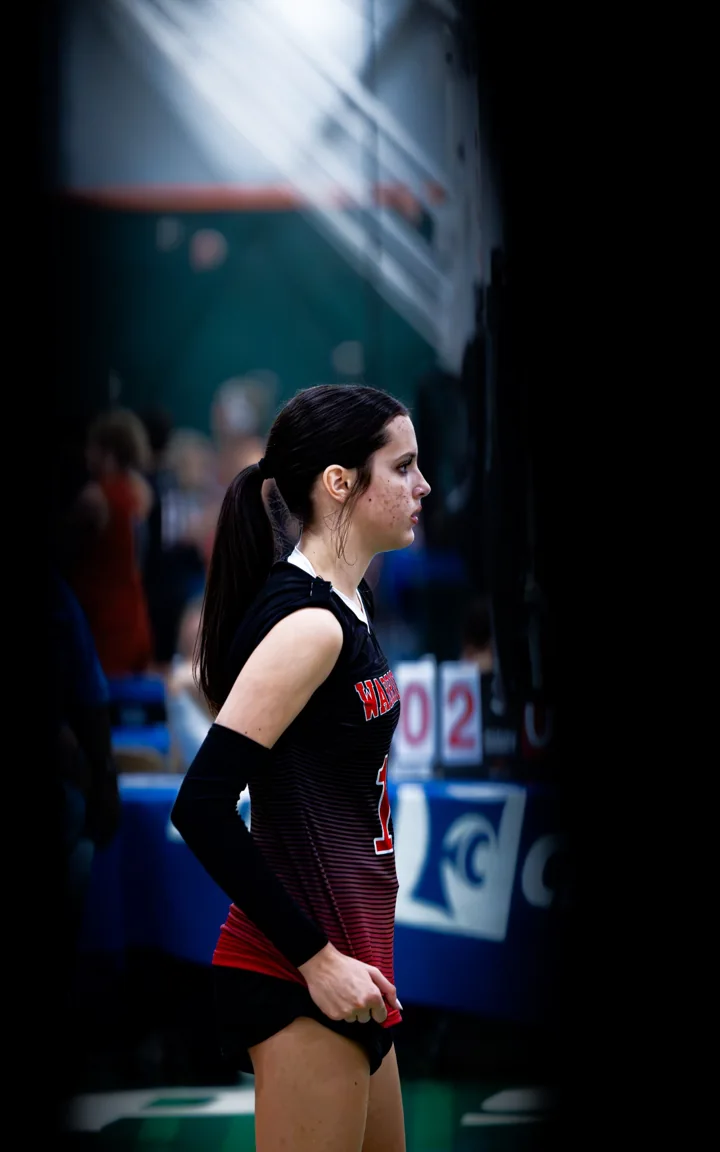 A volleyball player in red and black waits courtside, framed by the edge of the gym.