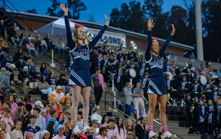 Cheerleaders perform elevated stunts in front of a crowded football grandstand.