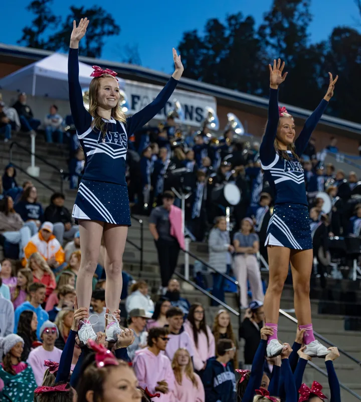 Cheerleaders perform elevated stunts in front of the football stands.