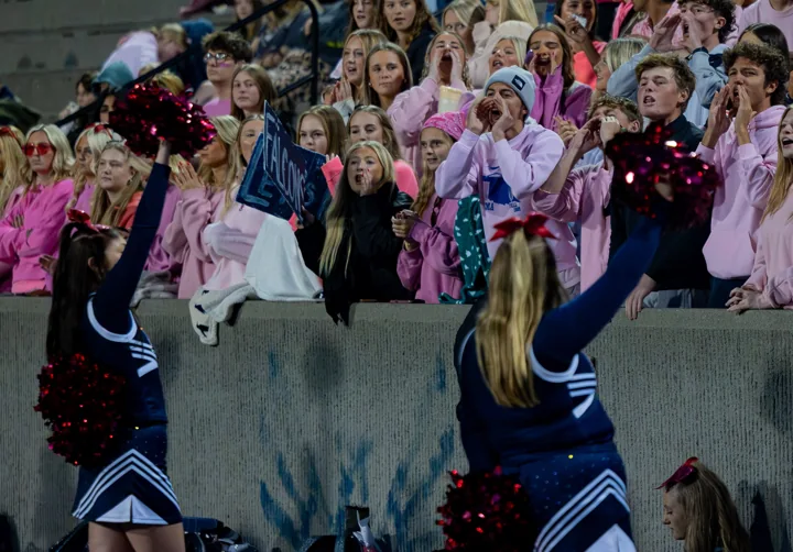 Students in the grandstand cheer while cheerleaders perform beside the field.