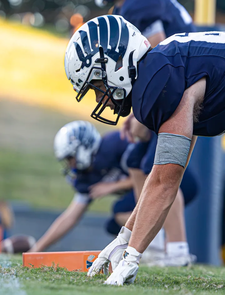 A football lineman leans forward over his hands with teammates lined up behind him.