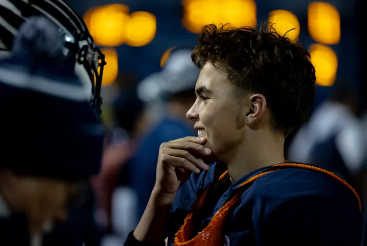 A football player in profile watches from the sideline, with a glowing scoreboard behind him.