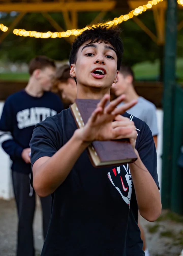 A young man gestures while holding a book outdoors beneath string lights.