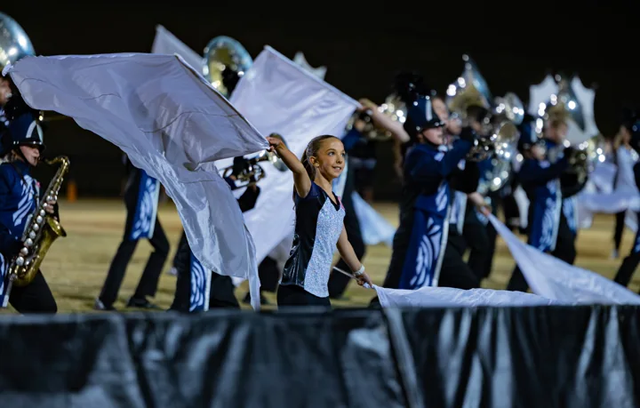 A color guard performer waves a pale flag with marching musicians behind her.