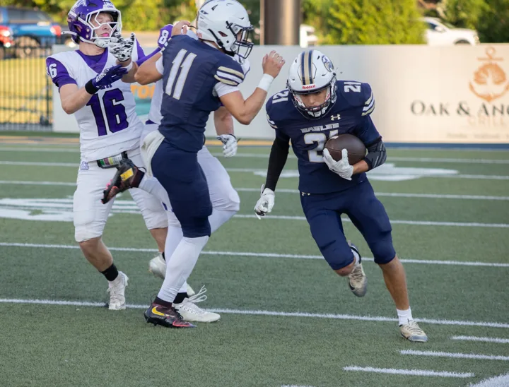 A football ball carrier in navy moves around a teammate as a defender approaches.