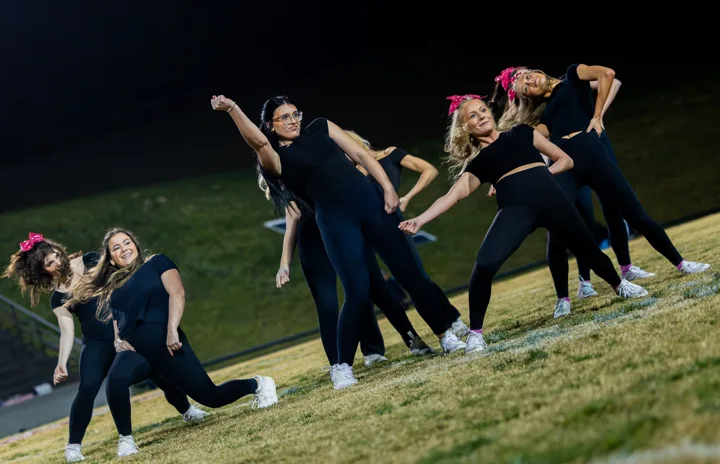 Performers in black strike coordinated poses on a field under evening lights.