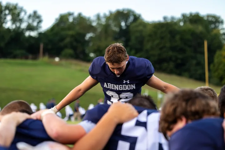 A football player leans into a team huddle with arms extended over his teammates.